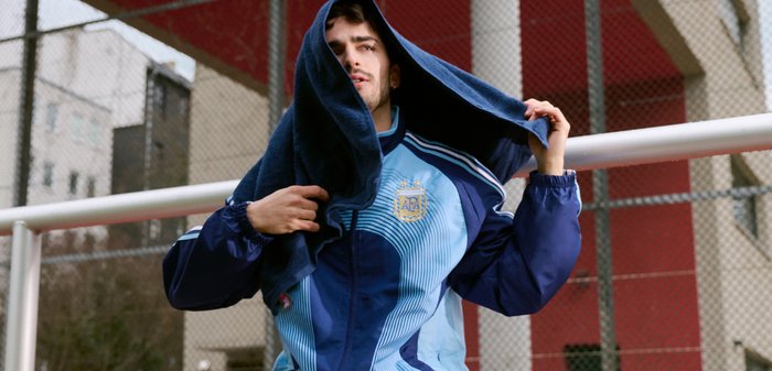 Jeune homme en veste de sport bleue soulevant une serviette sombre au-dessus de sa tête près d'une clôture en chaîne à côté d'un bâtiment rouge et blanc.