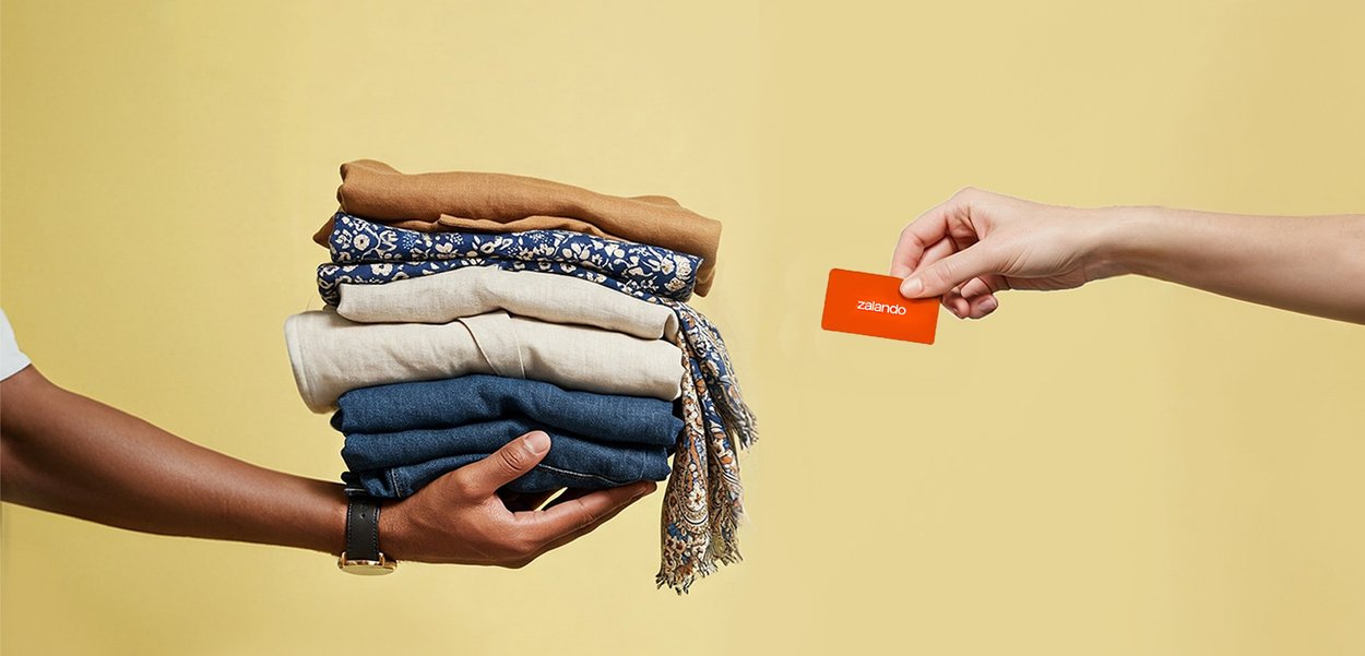 Hand holding folded clothes, other hand extending an orange Zalando card, beige background.
