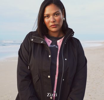 Veste noire à fermeture éclair avec une capuche, dotée de boutons-pression et de poches latérales, portée sur une chemise grise à pois roses et blancs.