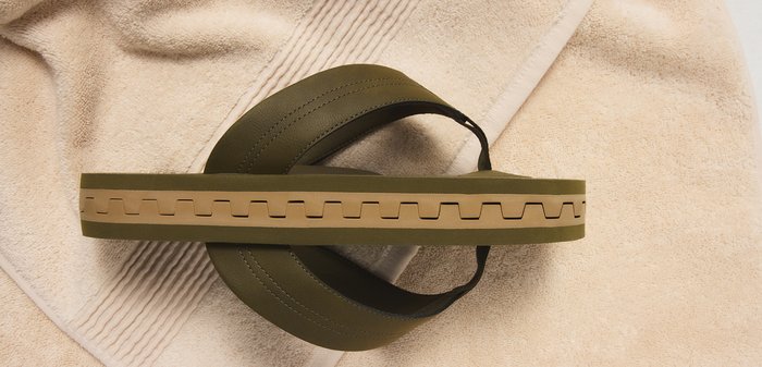 Olijfgroene en beige teenslipper met dikke zool, rustend op gestructureerde beige handdoeken.