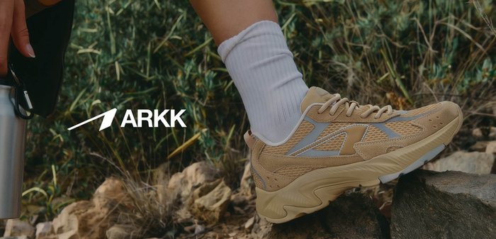 Person iført beige ARKK-sneakers og hvide sokker hviler foden på sten og holder en sølvfarvet vandflaske ude i det fri nær grønne områder.