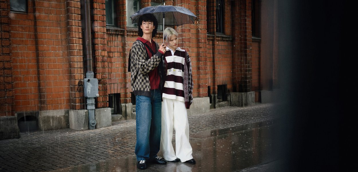 Două persoane stau sub o umbrelă neagră în ploaie. Una poartă un cardigan gri cu model peste un top roșu, blugi și pantofi negri. Cealaltă este îmbrăcată într-o cămașă cu dungi orizontale și pantaloni albi largi.