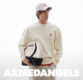 Uomo con felpa crema e berretto rosa tiene una borsa tote crema e nera coordinata, in piedi davanti a uno sfondo bianco con testo "ARMEDANGELS".