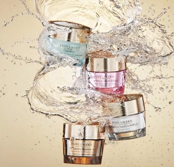 Fyra Estee Lauder-krämburkar i blått, rosa, brons och klart glas, med metalliska lock, som sprutas i vatten mot en neutral bakgrund.