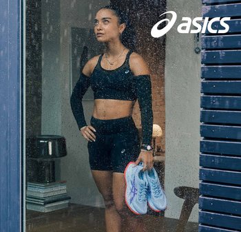 Zwarte atletische crop top en shorts; houdt blauwe hardloopschoenen vast met roze accenten. Regen op glas voegt textuur toe; binnenomgeving zichtbaar erachter.