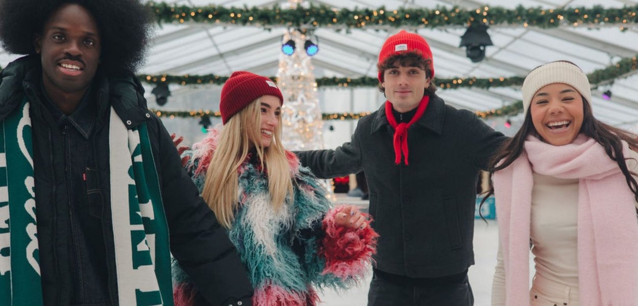 Vier mensen staan samen, gekleed in winterkleding: een zwarte donsjas, een kleurrijke, harige jas, een rode ski-muts en pastelkleurige sjaals.