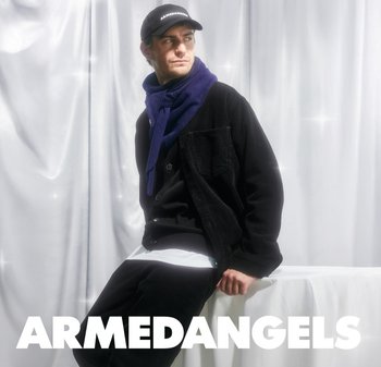 Black corduroy jacket over a white shirt, paired with black pants. A purple scarf and a black cap with "ARMEDANGELS" logo. Soft background.