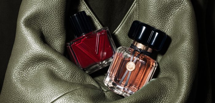 Deux flacons de parfum, un rouge et un ambré avec des bouchons noirs, reposant sur un tissu en cuir vert olive à texture douce.