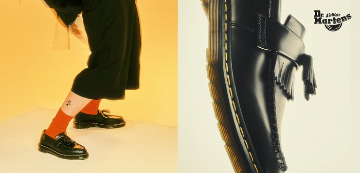 Mocassini in pelle nera con finitura testurizzata, con fibbia e dettaglio a frange, su sfondo giallo. Cuciture gialle che delineano la suola.