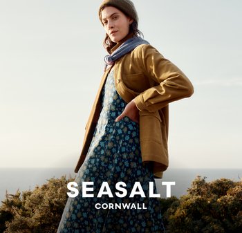 Žlutá manšestrová bunda přes modré květinové šaty, doplněná šedým šátkem a hnědým čepicí, na pozadí pobřežní krajiny.