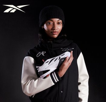 Frau mit schwarzer Mütze und Collegejacke hält einen weißen und schwarzen Sportschuh vor schwarzem Hintergrund, Reebok-Logo in der oberen Ecke.