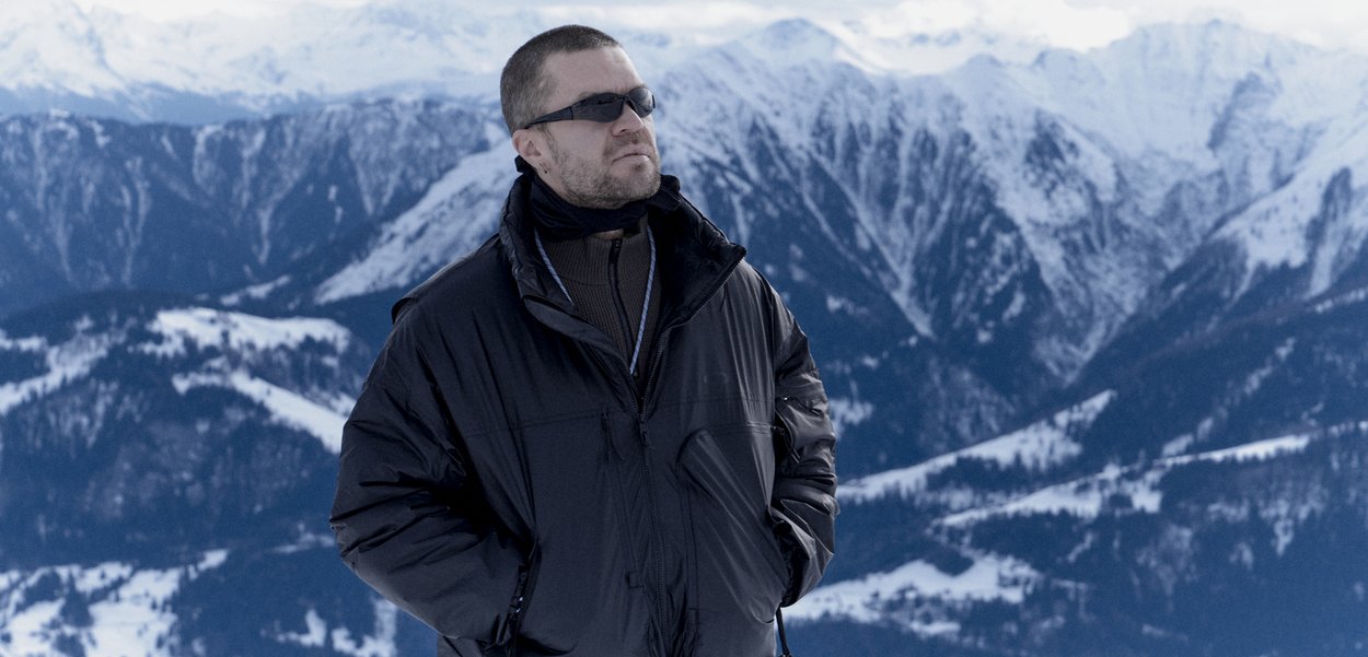 Mand iført solbriller og en sort vinterjakke står med hænderne i lommerne foran en sneklædt bjergryg.