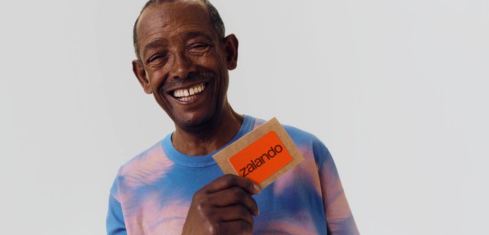 Homme souriant portant un t-shirt tie-dye bleu et rose, tenant une carte orange Zalando devant un fond clair uni.