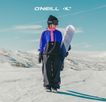 Snowboarder in een roze en blauwe kleurblokkering jas met zwarte broek, zwarte handschoenen en een zwarte beanie, die een snowboard vasthoudt in besneeuwd terrein.