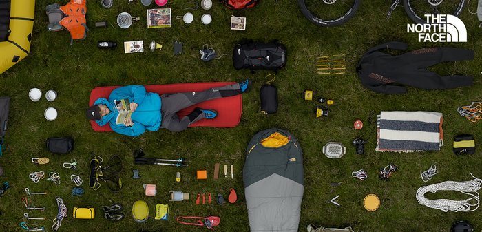 Camping-Ausrüstung, die auf Gras ausgebreitet ist, mit einem gelben aufblasbaren Schlauchboot, einem grauen Schlafsack sowie verschiedenen Utensilien, Seilen und Outdoor-Ausrüstung.