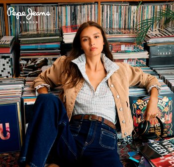 Chaqueta de cordones marrón con botones plateados, camisa blanca a cuadros, jeans azul oscuro, rodeado de discos de vinilo y coloridas portadas de álbumes.
