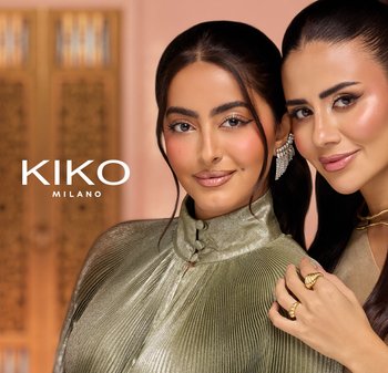 Dwie kobiety o gładkich ciemnych włosach mają na sobie elegancki makijaż i biżuterię, uśmiechając się blisko siebie na tle ciepłego, delikatnie rozmytego tła.