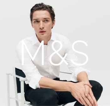 Chemise blanche à boutons, pantalon noir. Modèle assis, mains reposant sur les genoux, arrière-plan neutre. Gros logo "M&S" au centre.