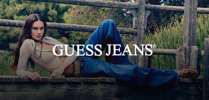 Młoda kobieta ubrana w beżową bluzę na suwak i niebieskie dżinsy stoi na drewnianym pomoście przy jeziorze, trzymając dziergane rękawiczki, otoczona drzewami.