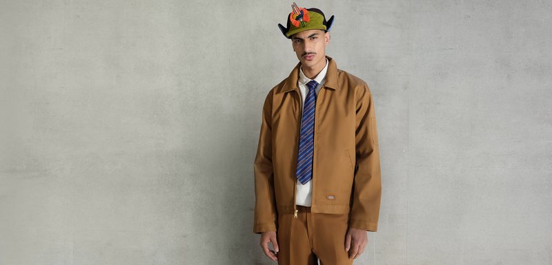 Homme portant une veste et un pantalon marron, une chemise blanche, une cravate rayée bleue et rouge, et un chapeau vert avec une décoration en plume colorée, se tenant contre un mur gris.
