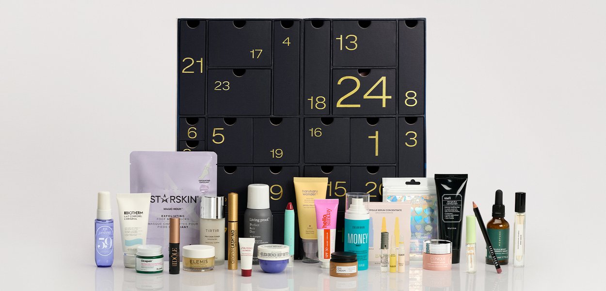 Julekalender med 24 sorte skuffer og forskellige hudpleje-, makeup- og skønhedsprodukter arrangeret foran på en reflekterende overflade.