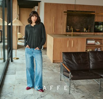 Grijze lange mouw top, lichtblauwe wijde jeans en bordeauxrode schoenen. Moderne keukensetting met houten kasten en marmeren vloeren.