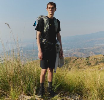 Jeune homme debout sur une colline herbeuse, vêtu d'un équipement de randonnée et portant un sac à dos, avec des montagnes visibles au loin.