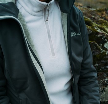 Dunkelgraue Jacke mit Kapuze und hellgrauem Fleece-Futter, über einem weißen Fleece-Oberteil mit Reißverschluss getragen. Das Markenlogo ist auf der Jacke sichtbar.