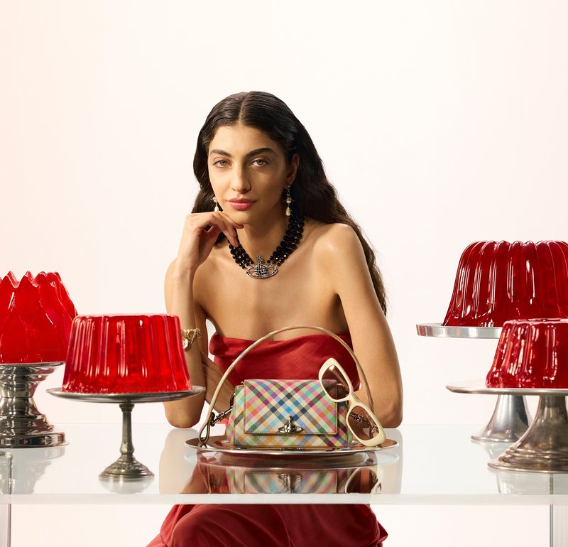 Femme en robe rouge bustier et collier noir à perles assise derrière une table transparente avec un sac à main coloré, des lunettes de soleil et des desserts en gelée rouges sur des présentoirs.