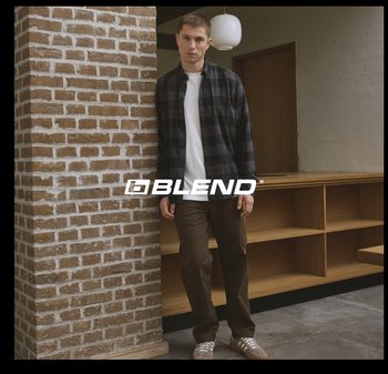 Casual outfit met een geruit zwart-wit-grijs overhemd over een wit T-shirt, gecombineerd met bruine broek en grijze sneakers, tegen een bakstenen muur.