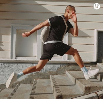Maglietta sportiva nera con accenti bianchi, pantaloni corti neri, scarpe da ginnastica bianche e calze azzurro chiaro. Sta correndo su gradini di cemento, portando uno zainetto leggero.