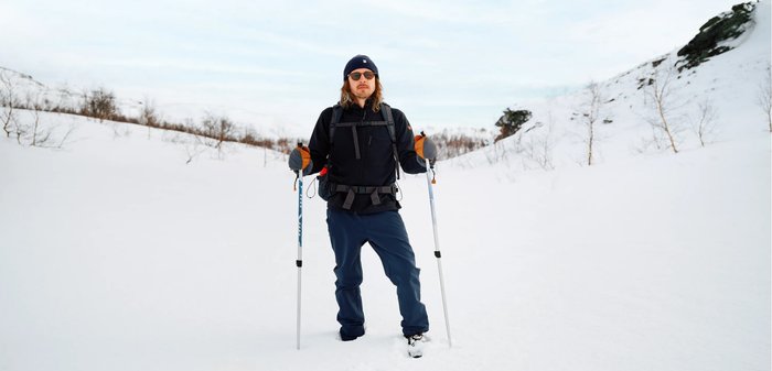 Eine Person in einer schwarzen Jacke und blauen Hosen steht im Schnee. Sie hält Trekkingstöcke und trägt Handschuhe, eine Mütze und Sonnenbrille.