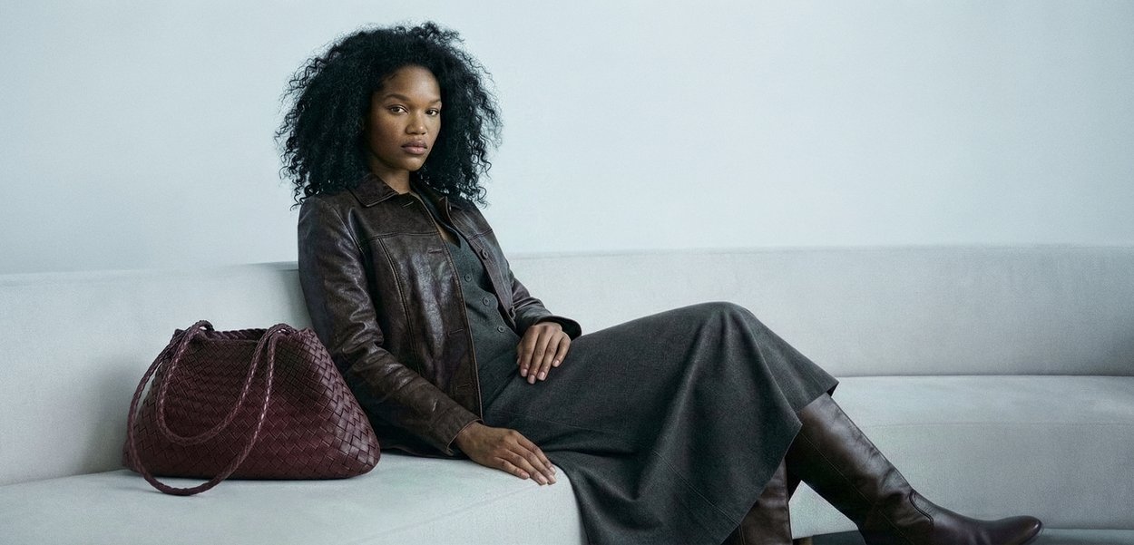 Femeie cu păr creț stând pe o canapea gri deschis, purtând o jachetă maro din piele, o rochie gri și cizme maro, cu o geantă mare maro împletită.