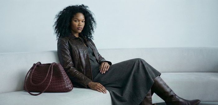 Γυναίκα με κυματιστά μαλλιά που κάθεται σε ένα ανοιχτό γκρι καναπέ, φορώντας ένα καφέ δερμάτινο μπουφάν, γκρι φόρεμα και καφέ μπότες, με μια μεγάλη πλεκτή καφέ τσάντα.