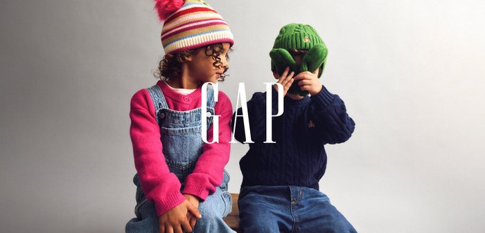 To børn sidder side om side. Den ene bærer en lyserød sweater og denimoveralls; den anden har en mørkegrøn hue og en marineblå kabelstrikket sweater.