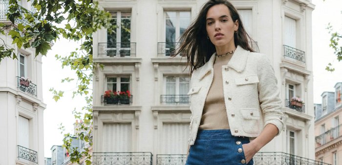 Ung kvinna med långt mörkt hår bär en vit strukturerad jacka med knappar och blå byxor med hög midja, stående på en stadsgata.