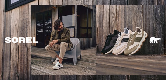 Mann sitzt auf einem Stuhl auf einem hölzernen Deck und trägt Freizeitschuhe; drei Paare Freizeitsneaker sind in der Nähe auf einer Holzoberfläche ausgestellt.