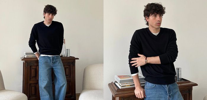 Fiatal férfi göndör hajjal, fekete pulóvert és kék farmert viselve, állva pózol egy fa szekrény mellett, amelyben könyvek és egy fém pohár található.