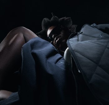 Persona con capelli strutturati e occhiali da sole, reclinata indossando una giacca trapuntata scura e tessuto strutturato su sfondo nero.