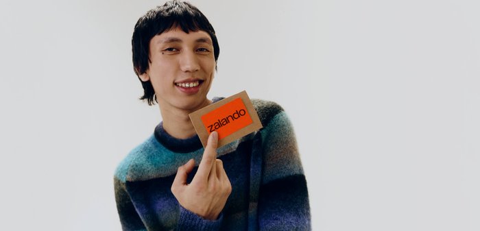 Giovane sorridente che tiene in mano una carta regalo Zalando su uno sfondo chiaro uniforme.