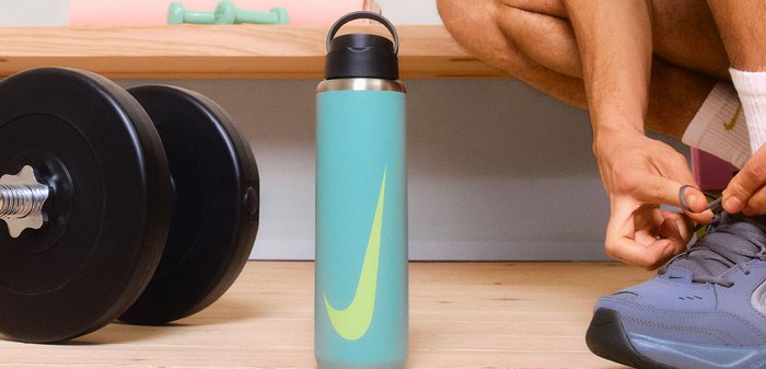 Πράσινο παγούρι από ανοξείδωτο χάλυβα με μεγάλο μαύρο καπάκι και κίτρινο λογότυπο. Μαύρα αλτήρες στο προσκήνιο, ξύλινη επιφάνεια από κάτω.