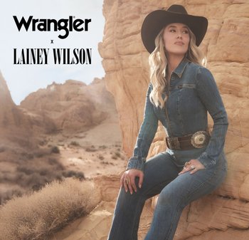 Denim jumpsuit with long sleeves and fitted style, featuring a wide leather belt with a large round buckle, set against a sandy landscape.