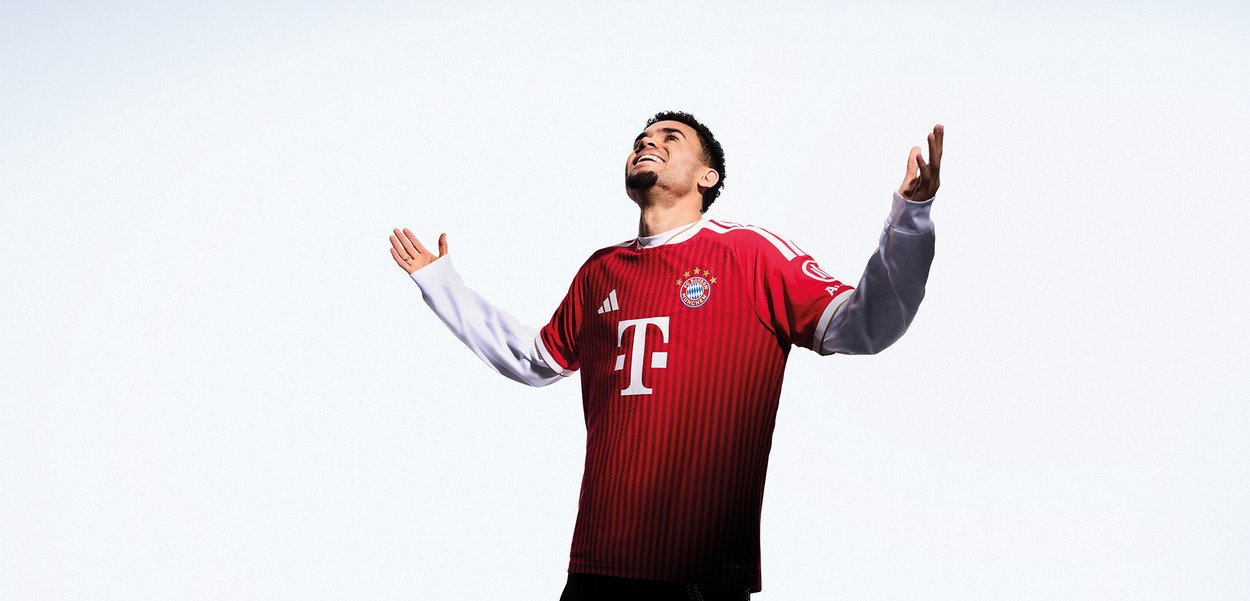 Man wearing Bayern Munich football jersey with arms raised, looking upward and smiling against a plain white background.