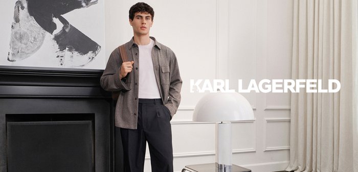 Man in grey shirt and black trousers stands by a fireplace, holding a backpack strap. Modern lamp and abstract art are in the background.