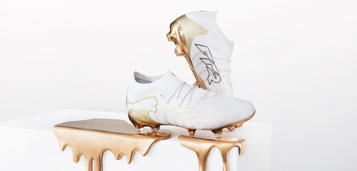 Chaussures de football blanches avec des accents dorés, surface texturée et design distinctif de la semelle, reposant sur un socle blanc épuré avec un effet de coulure dorée.