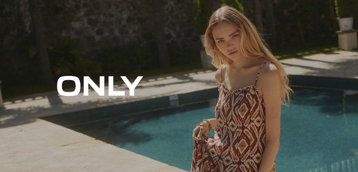 Mulher loira com um vestido de verão estampado está perto de uma piscina com uma parede de pedra e vegetação ao fundo.