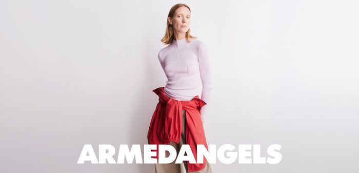 Woman in a light pink sweater with a red jacket tied around her waist, standing against a white background with "ARMEDANGELS" text below.