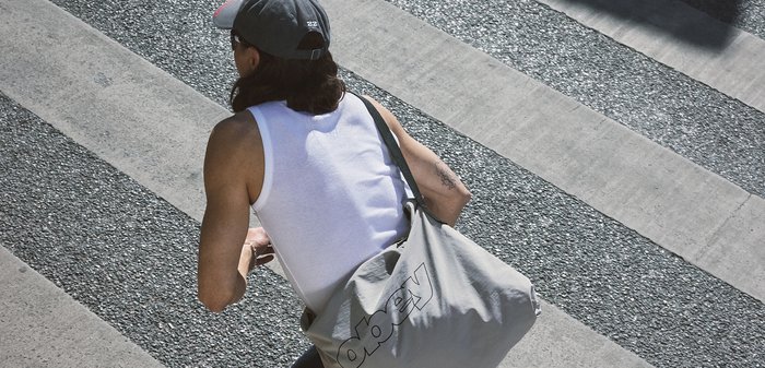 Άτομο που φοράει λευκό αμάνικο και καπέλο, κρατώντας μια μεγάλη γκρι τσάντα Obey, περπατώντας πάνω σε υφασμένη διάβαση πεζών.