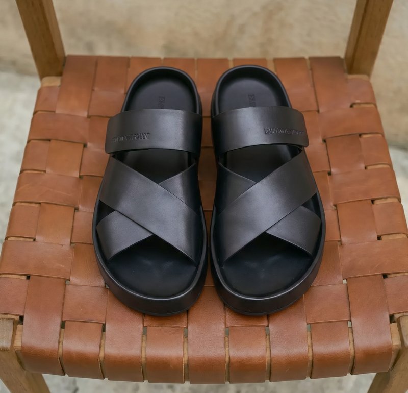 Ζευγάρι μαύρα δερμάτινα σανδάλια με σταυρωτούς ιμάντες τοποθετημένα σε καφέ πλεκτή δερμάτινη καρέκλα με ουδέτερο φόντο.