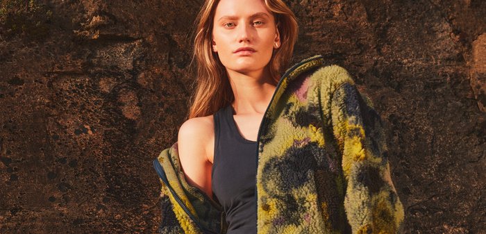 Tânără cu părul lung blond, purtând o jachetă din polar cu model floral peste o bluză fără mâneci neagră și pantaloni largi închiși la culoare, în fața unui perete de piatră texturat.